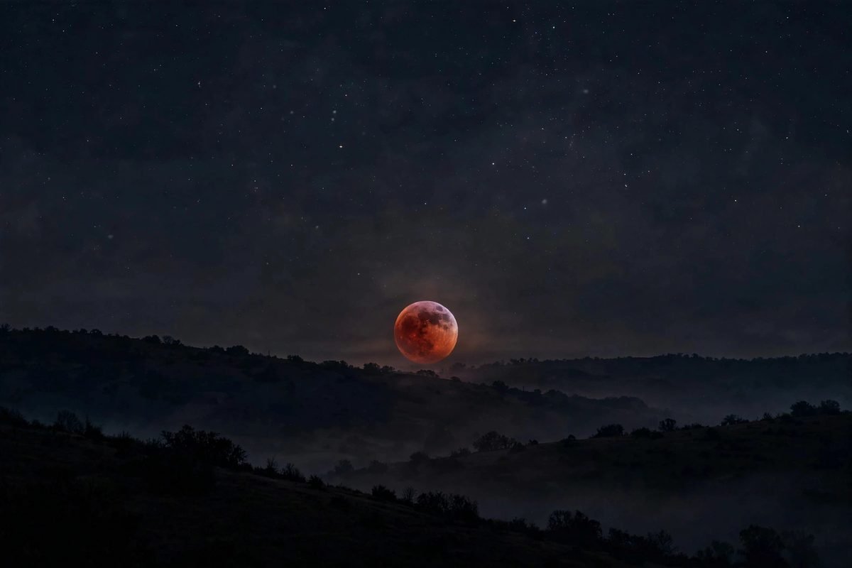 A copper-red blood moon low over a dark hillside during totality.